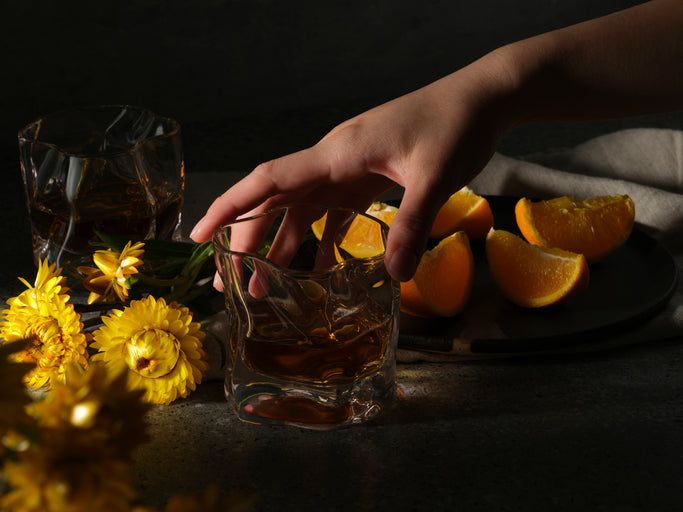 A hand reaches for a Malt & Brew Whisky Wave glass, surrounded by flowers and fruit in chiaroscuro.