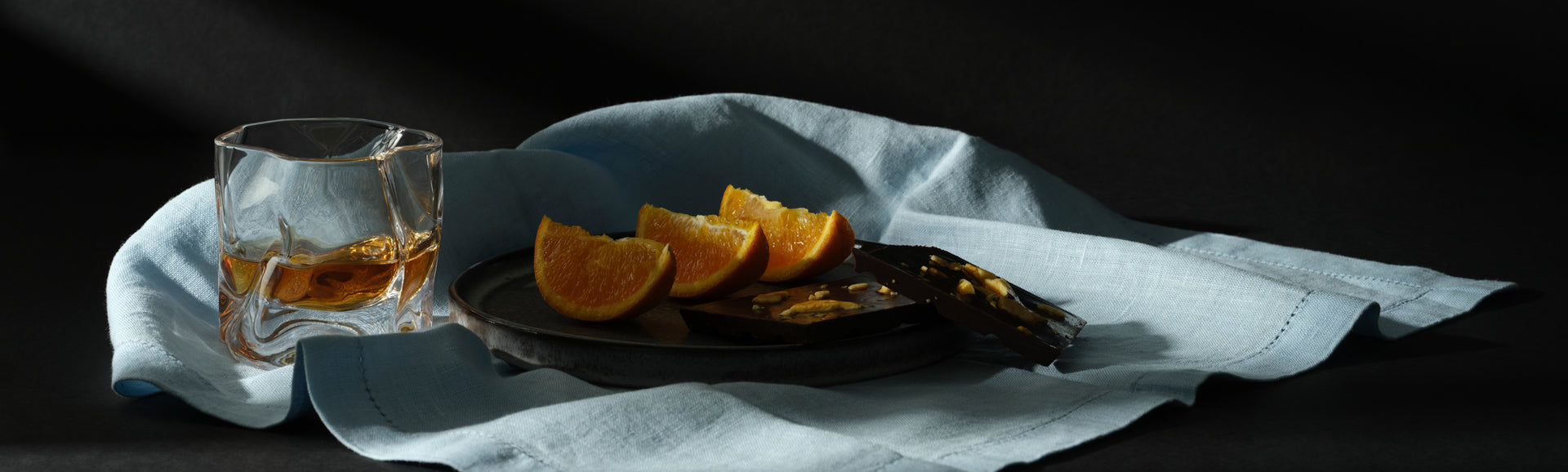 A Malt & Brew Whisky Wave Glass sits on a blue cloth with a plate of orange slices and dark chocolate.