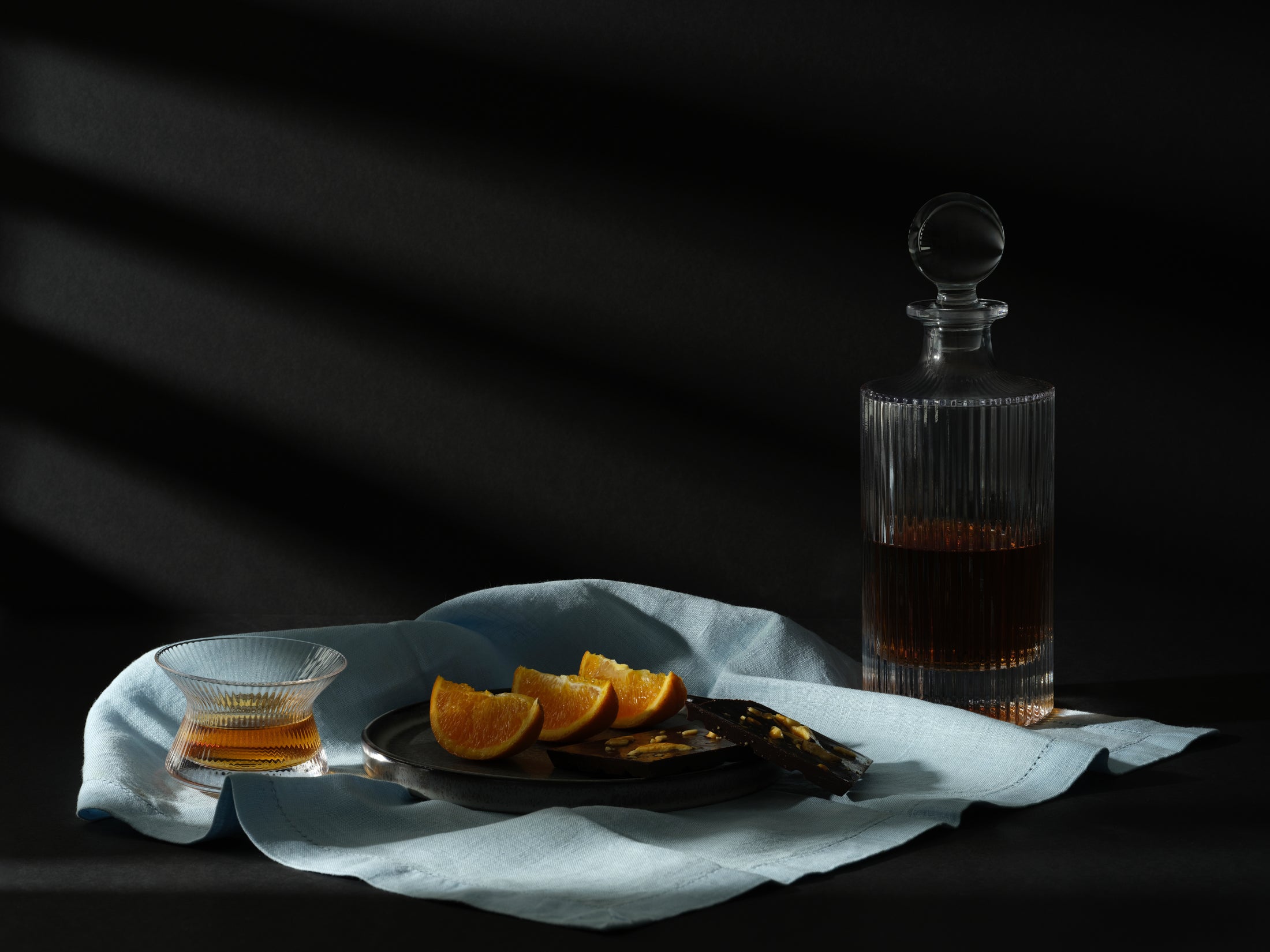 A Malt & Brew Hanyu Glass sits on a blue cloth with oranges, dark chocolate, and a whisky decanter.