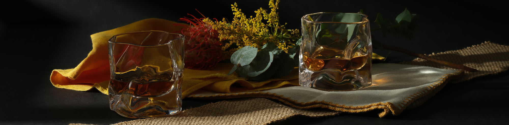 Two Malt & Brew Whisky Wave glasses sit among yellow flowers and fabrics.