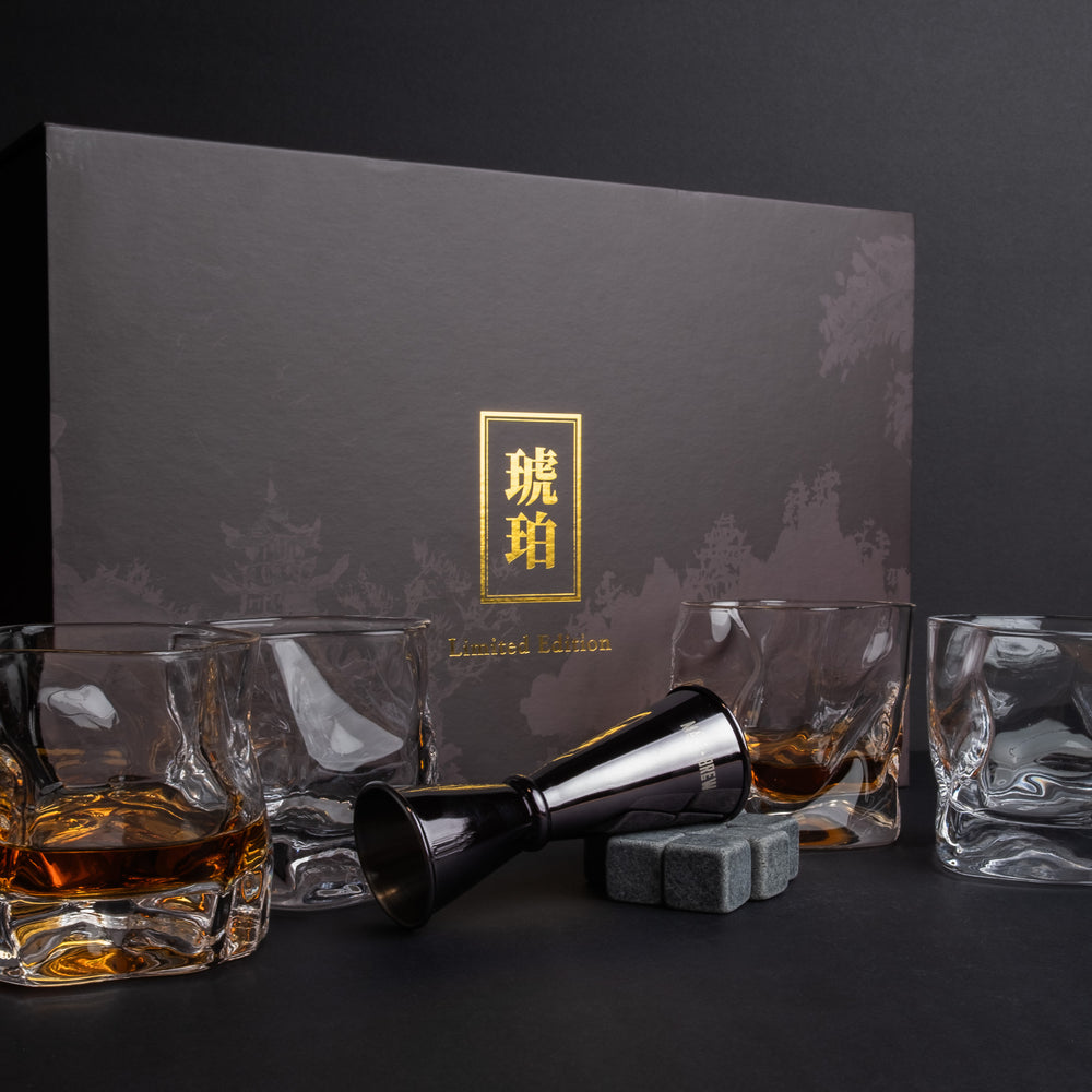 A whiskey glass set with a dark gray presentation box, including four clear whiskey glasses, a black jigger, and two gray ice cubes, arranged on a dark surface with a dark background.