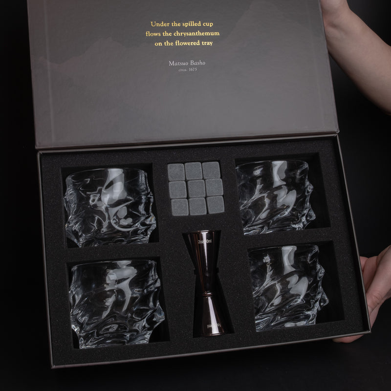 A premium crystal whiskey glass set in an open presentation box, featuring four textured clear crystal glasses, a central metallic jigger, and four gray ice cubes. The dark gray box lid has gold text and is held open by hands at the top right and bottom right.